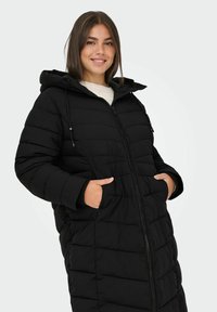 Abrigo puff negro con capucha, con cierre frontal, dos bolsillos laterales y diseño acolchado. Textura suave y aislamiento cálido.