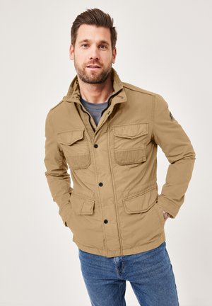 Beige, textured utility jacket with four front pockets, a zippered closure, and a stand-up collar, paired with blue jeans.