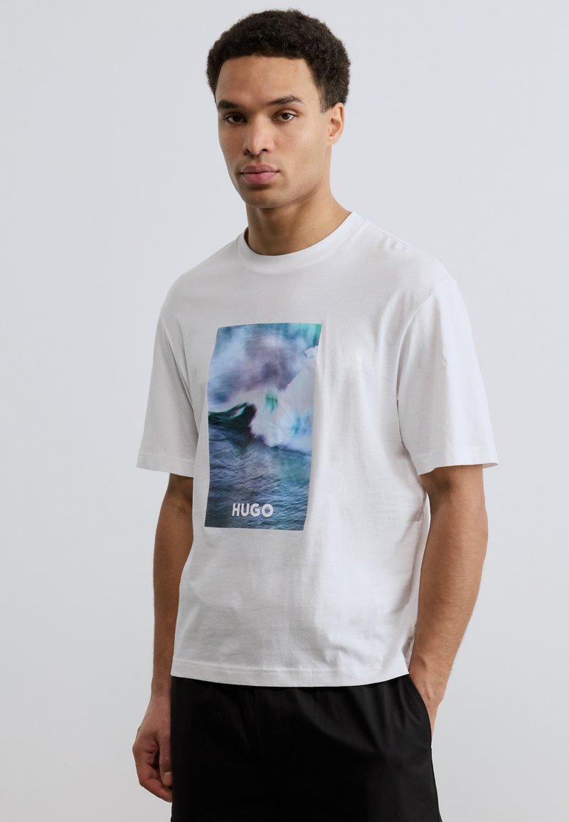 Νέος άνδρας που φοράει λευκό T-shirt με μπλε γραφικό κυματιστού ωκεανού και κείμενο "HUGO", συνδυασμένο με μαύρο παντελόνι, στέκεται μπροστά σε απλό φόντο.
