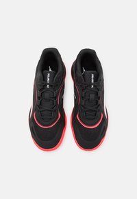 Zapatillas deportivas negras con acentos rojos; materiales de malla y sintéticos; puntera redondeada; cordones y lengüeta con marca. La suela tiene un patrón texturizado y antideslizante.