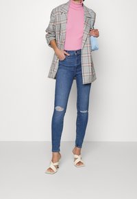 Blazer a cuadros, suéter de cuello alto rosa, jeans azul desgastados, sandalias de tacón color crema y un bolso azul. Incluye mangas arremangadas y desgarros en los jeans.