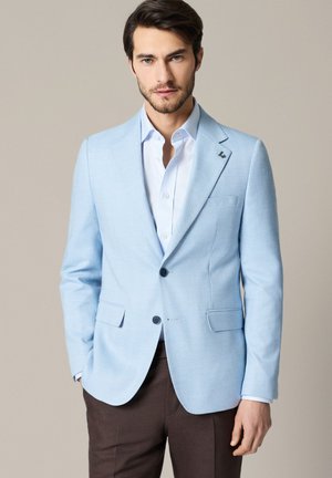 Mężczyzna w jasnoniebieskiej marynarce na białej koszuli i ciemnobrązowych spodniach na jednolitym tle, ręce w kieszeniach.