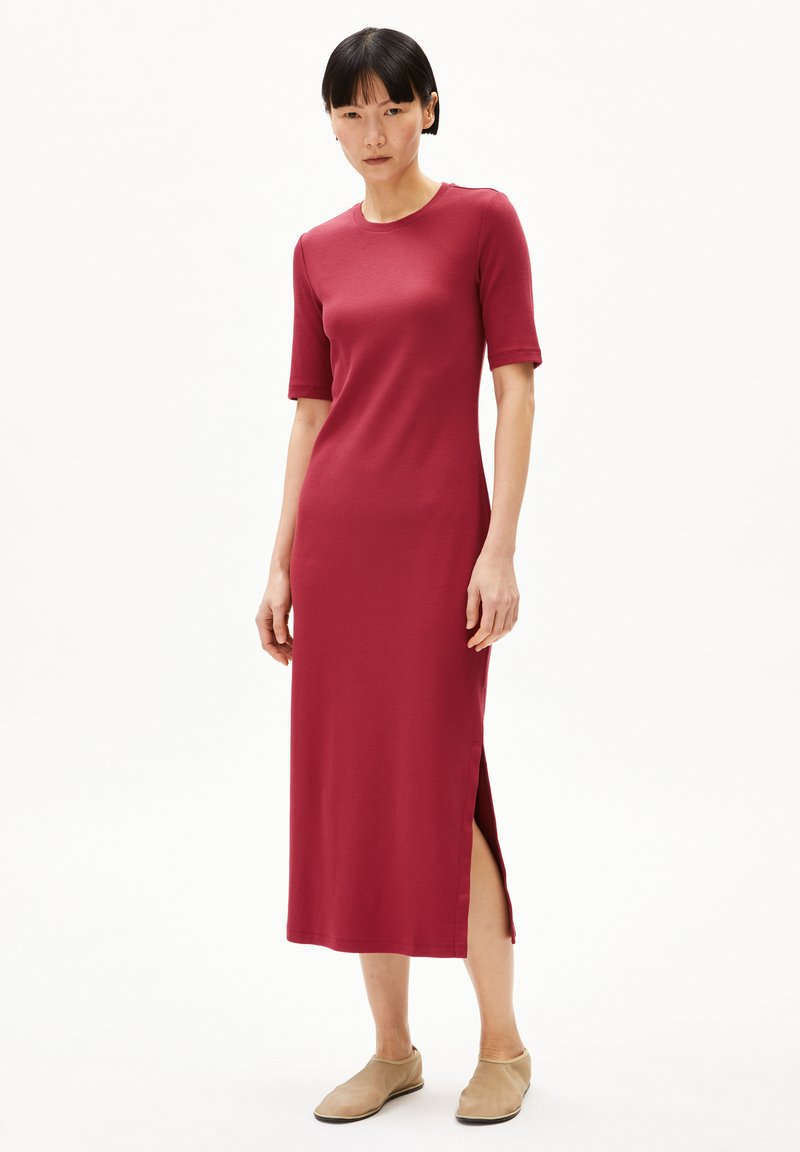 Geribbelde, knielange jurk in diep rood met korte mouwen en zijsplitten, gecombineerd met beige instappers. Soepele textuur, fitted ontwerp.