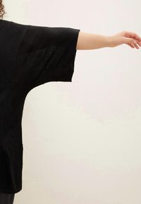 Czarna, luźna bluzka z szerokimi rękawami, wykonana z przezroczystego materiału z fakturowanymi wzorami. Ręka wyciągnięta, prezentująca krój odzieży.