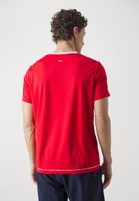 Camiseta atlética roja de manga corta, con cuello redondo y ribete blanco en el cuello y el dobladillo. Presenta un logo en la parte superior de la espalda. Textura suave.