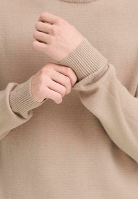 Beiger Pullover mit gerippten Bündchen und rundem Halsausschnitt, featuring eine weiche Stricktextur und Nahaufnahme von Händen, die die Ärmel anpassen.