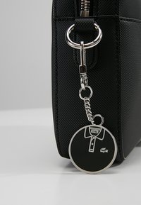 Czarna torebka o teksturowej powierzchni z srebrnym zamkiem błyskawicznym, z okrągłym brelokiem w kształcie koszulki oraz małym zielonym logo.