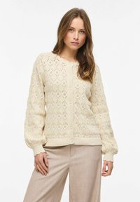 Femme portant un cardigan en maille crème boutonné à manches longues avec un motif en losanges et un pantalon beige, debout devant un fond uni.