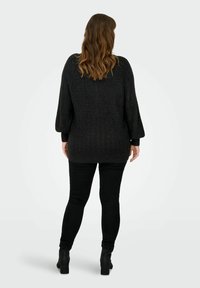 Čierny pletený sveter s textúrovaným vzorom, vo voľnom strihu a trojštvrťovými rukávmi, v kombinácii s čiernymi priliehavými nohavicami a členkovými čižmami.