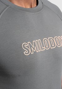 Harmaa puuvillapaita, jossa on pyöreä kaula-aukko ja edessä suuri tekstilogo "SMILODONY" vaalean persikan värissä. Liukas tekstuuri, istuva muotoilu.