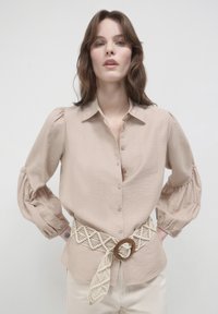 Camicia beige con bottoni, maniche a sbuffo e finitura testurizzata, abbinata a una cintura di crema intrecciata con una fibbia circolare in legno.