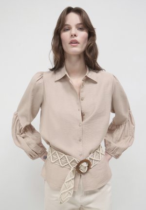 Camicia beige con bottoni, maniche a sbuffo e finitura testurizzata, abbinata a una cintura di crema intrecciata con una fibbia circolare in legno.