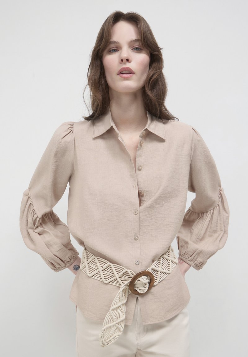 Camicia beige con bottoni, maniche a sbuffo e finitura testurizzata, abbinata a una cintura di crema intrecciata con una fibbia circolare in legno.