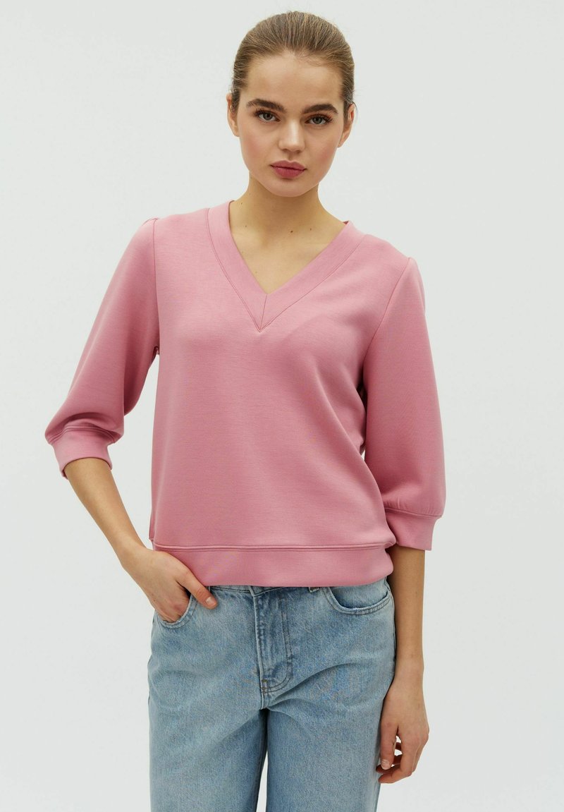 Jeune femme portant un sweat-shirt rose à col en V avec des manches trois-quarts et un jean bleu clair, debout devant un fond clair uni.