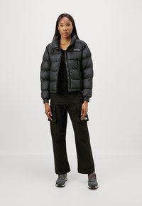 Geacă puffer neagră cu guler înalt, închidere cu fermoar și manșete elastice, purtată cu pantaloni cargo negri și încălțăminte gri de drumeție.