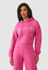 Różowa bluza z kapturem wykonana z miękkiego materiału, z kieszenią z przodu, sznurkami i detalami z kryształków w logo. Stylizowana z pasującymi różowymi spodniami.