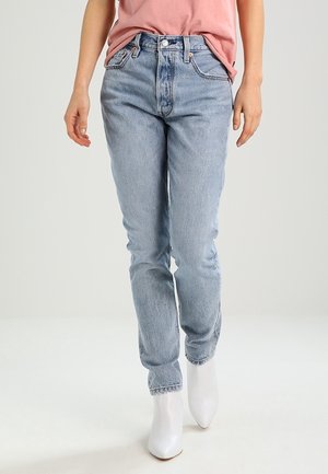 Person iført lyseblå jeans, en lyserød skjorte stukket ned i bukserne, og hvide spidse ankelsko, der står mod en ensfarvet baggrund.