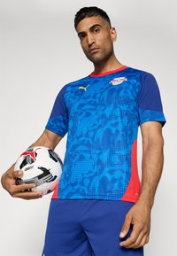 Maglietta sportiva a maniche corte blu e rossa con un motivo geometrico testurizzato. Il giocatore tiene un pallone da calcio bianco e nero con accenti rossi.