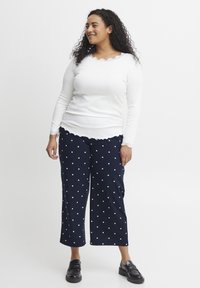 Biely dlhý rukáv s výstrihom do vlnky spárovaný s námorníckymi cropped nohavicami s bielymi bodkami. Modelka má na sebe čierne mokasíny.