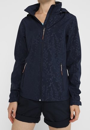 Frau, die eine marineblaue Jacke mit Reißverschluss und dezenten Blattmustern sowie schwarze hochgekrempelte Shorts trägt, gezeigt vom Hals bis zur Mitte des Oberschenkels.