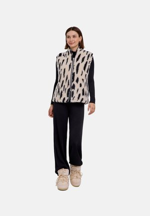 Frau trägt ein schwarzes Langarmoberteil, weite schwarze Hose, beige Stiefel und eine weiße Fleeceweste mit schwarzen abstrakten Flecken, steht vor weißem Hintergrund.