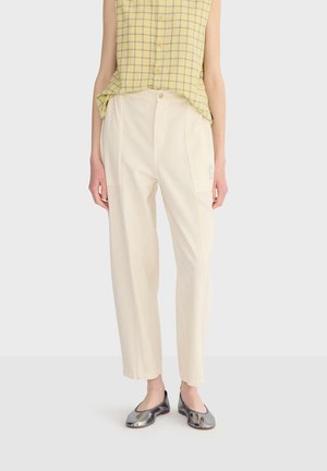 Femme portant un pantalon crème taille haute avec poches appliquées, un haut sans manches à carreaux jaunes, et des ballerines argentées métalliques, debout devant un fond blanc.