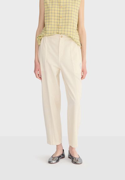 Femme portant un pantalon crème taille haute avec poches appliquées, un haut sans manches à carreaux jaunes, et des ballerines argentées métalliques, debout devant un fond blanc.