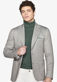 Lys grå hørfrakke med fiskebensmønster og to forlommer, båret over en mørkegrøn ribbet turtleneck. Hvid lommetørklæde med grønne accenter.