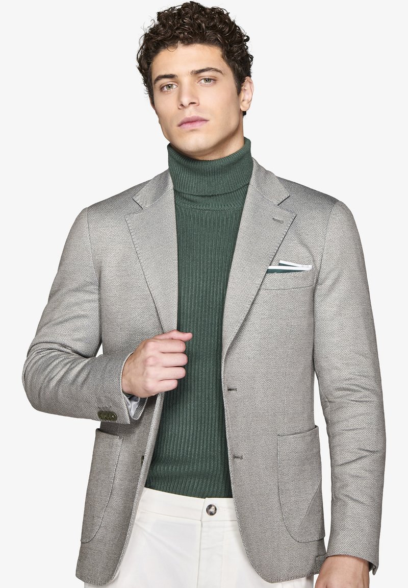 Ljust grå herringbone-blazer med två framfickor, bärs över en mörkgrön ribbad polotröja. Vit ficknäsa med gröna accenter.