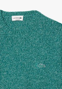 Maglione lavorato a maglia turchese con scollatura rotonda. Presenta un motivo testurizzato e un piccolo logo di coccodrillo ricamato sul davanti.