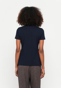 Marineblaue Kurzarm-T-Shirt mit Rundhalsausschnitt. Glatter Stoff, taillierte Form. Kombiniert mit gemusterten braunen Hosen, von hinten betrachtet.