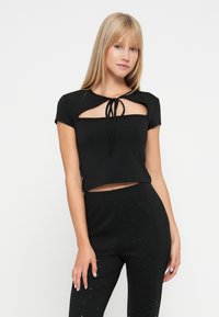 Mladá žena s rovnými blond vlasmi, oblečená v čiernom crop top s výrezmi a čiernych nohaviciach s vysokým pásom s jemnými bodkami, stojaca pred bielym pozadím.