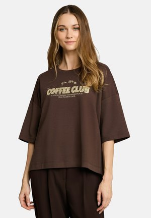 Jonge vrouw met lang lichtbruin haar draagt een loszittend bruin T-shirt met de tekst "Coffee Club" en bijpassende bruine broek tegen een effen achtergrond.