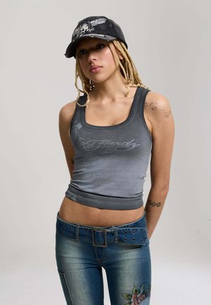 Giovane donna con capelli biondi intrecciati che indossa un berretto nero, una canotta grigia e jeans blu con ricami floreali e una cintura larga.