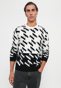 Junger Mann mit kurzen Haaren trägt einen schwarz-weißen, geometrisch gemusterten Pullover und dunkelgraue Jeans, steht vor einem schlichten weißen Hintergrund.
