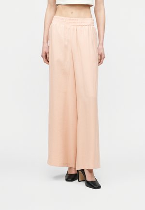 Kvinde iført lyserøde vidde bukser med elastisk talje og sorte blokhælte sko med firkantet tå, stående mod ensfarvet baggrund.