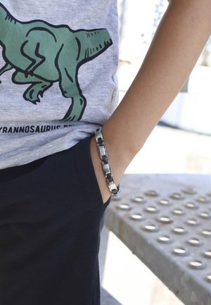 Metalen armband met verbonden schakels en een glanzende afwerking, gedragen om de pols van een kind, gecombineerd met een grijze t-shirt en donkere broek.