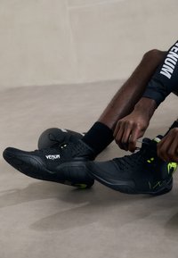 Black athletic shoes with a textured upper, neon green accents, and a sturdy sole. The laces are being tightened on the right shoe.