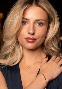 Gold necklace with a small gem pendant, worn with a gold bangle. Subject has long blonde hair and is dressed in a dark-colored top.