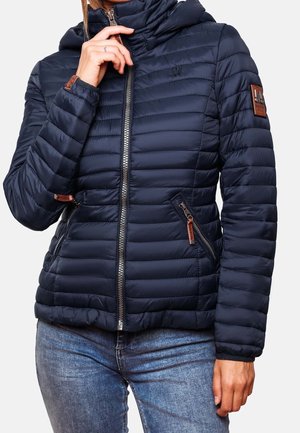 Personne portant une doudoune matelassée bleu foncé avec des poches zippées et un col montant, associée à un jean en denim bleu.