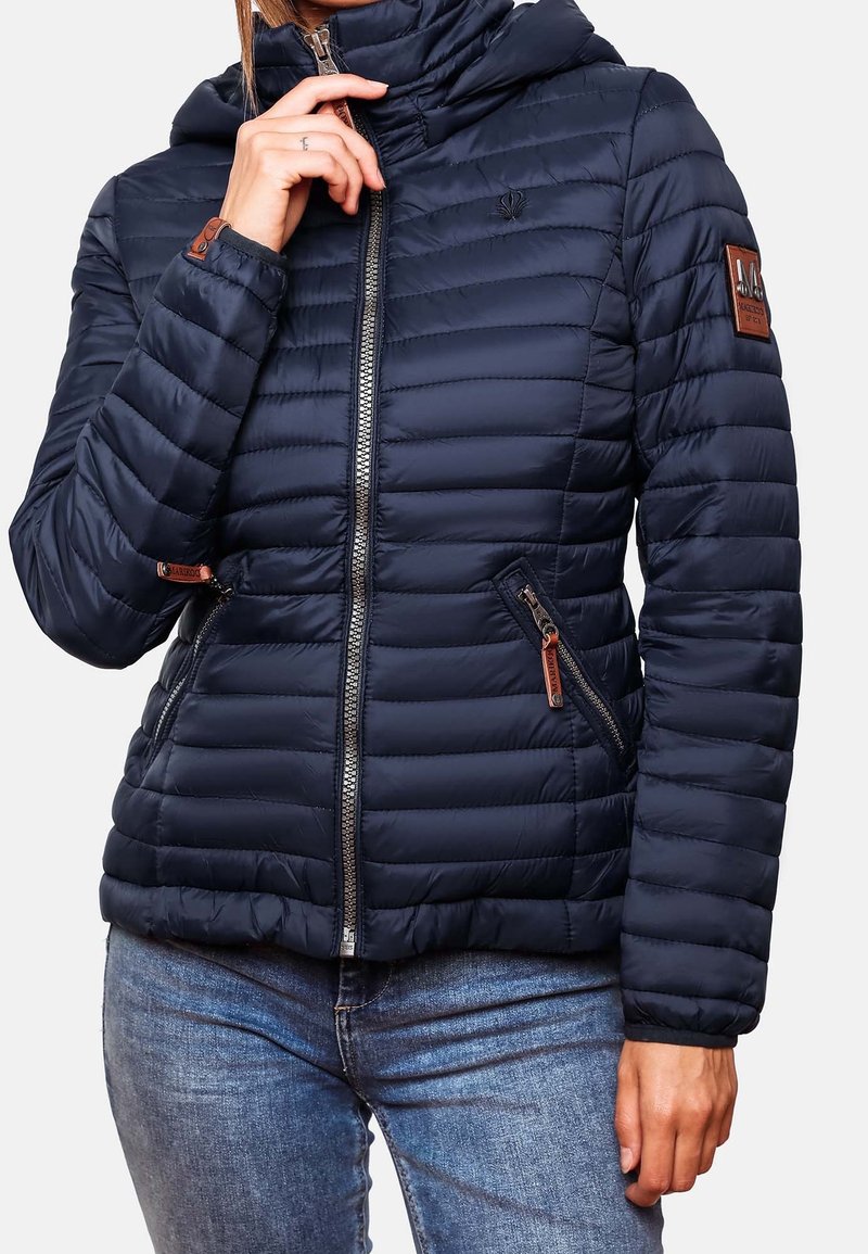 Personne portant une doudoune matelassée bleu foncé avec des poches zippées et un col montant, associée à un jean en denim bleu.