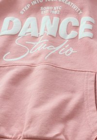Różowy sweatshirt z napisem „DANCE STUDIO” w białych blokowych literach oraz kursywą. Teksturowana tkanina z kieszenią kangurką z przodu.