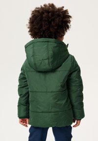 Grüne gepolsterte Jacke mit Kapuze, mit gestuften Bereichen und glatter Textur. Getragen mit marineblauen Hosen.