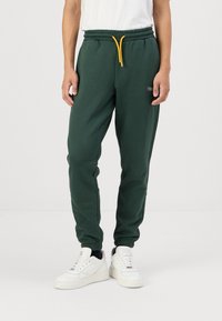 Green sweatpants made of soft fabric, featuring an elastic waistband with a yellow drawstring and cuffed hems. Simple embroidered logo detail.