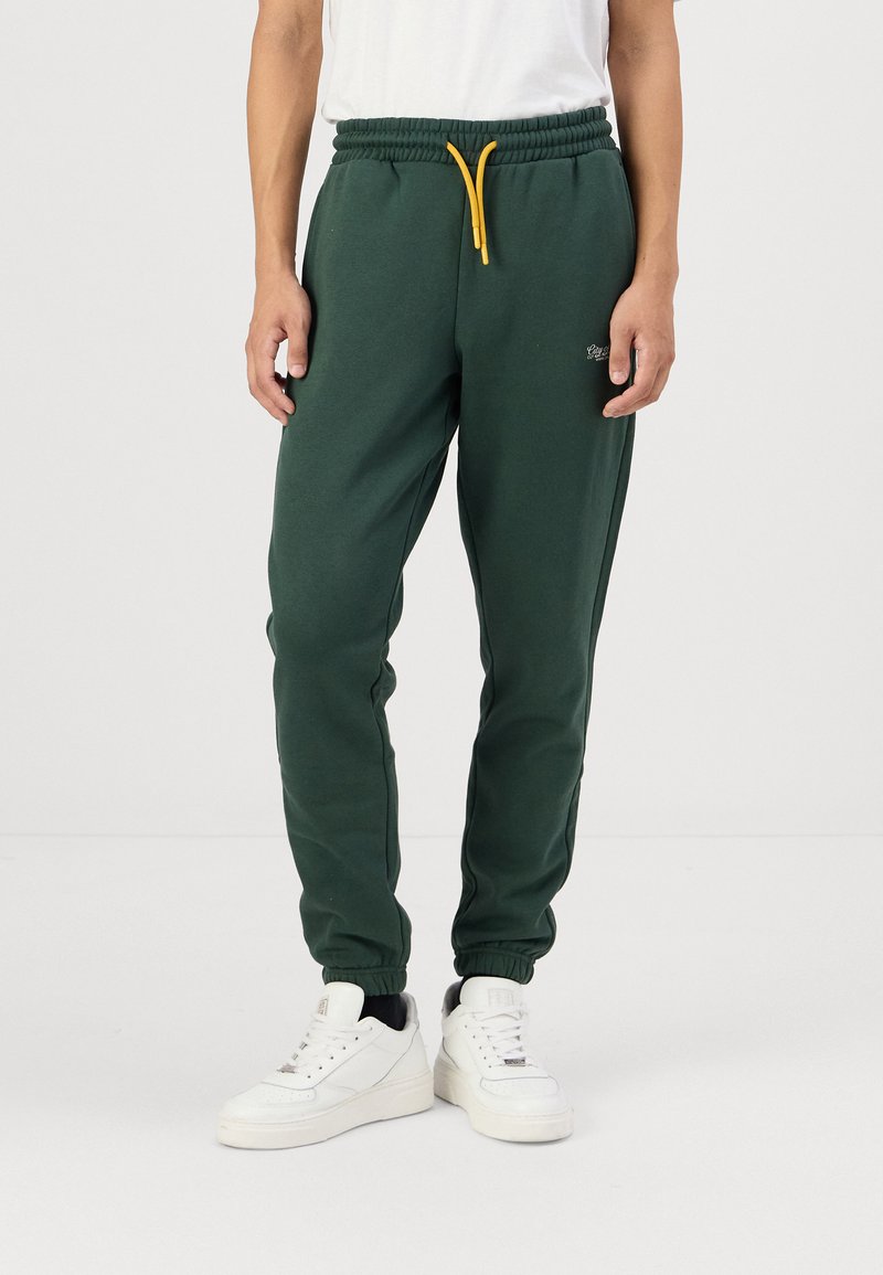 Green sweatpants made of soft fabric, featuring an elastic waistband with a yellow drawstring and cuffed hems. Simple embroidered logo detail.