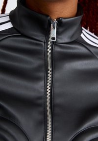 Primer plano de una chaqueta de cuero negra con una cremallera plateada que presenta el logo de Adidas, usada con detalle de rayas blancas en los hombros.