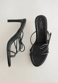 Zwarte high-heeled sandalen met slanke banden en strikken, met een gladde textuur en een gestroomlijnd ontwerp. Merkennaam "NEXT" zichtbaar op de binnenzool.