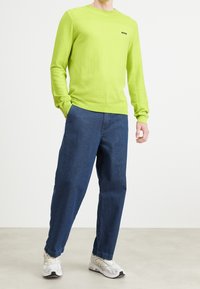 Man som bär en limegrön långärmad tröja, blå jeans och vita sneakers, står med ena handen i fickan på en neutral bakgrund.