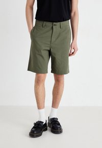 Shorts verts olive avec une coupe droite, deux poches avant et une fermeture à bouton. Associés à des mocassins noirs et des chaussettes blanches.