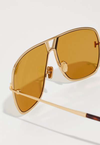 Gouden metalen aviator zonnebril met bruine lenzen, met een gedurfde hoekige vorm, plastic tempelpunten en een dubbele brugconstructie.
